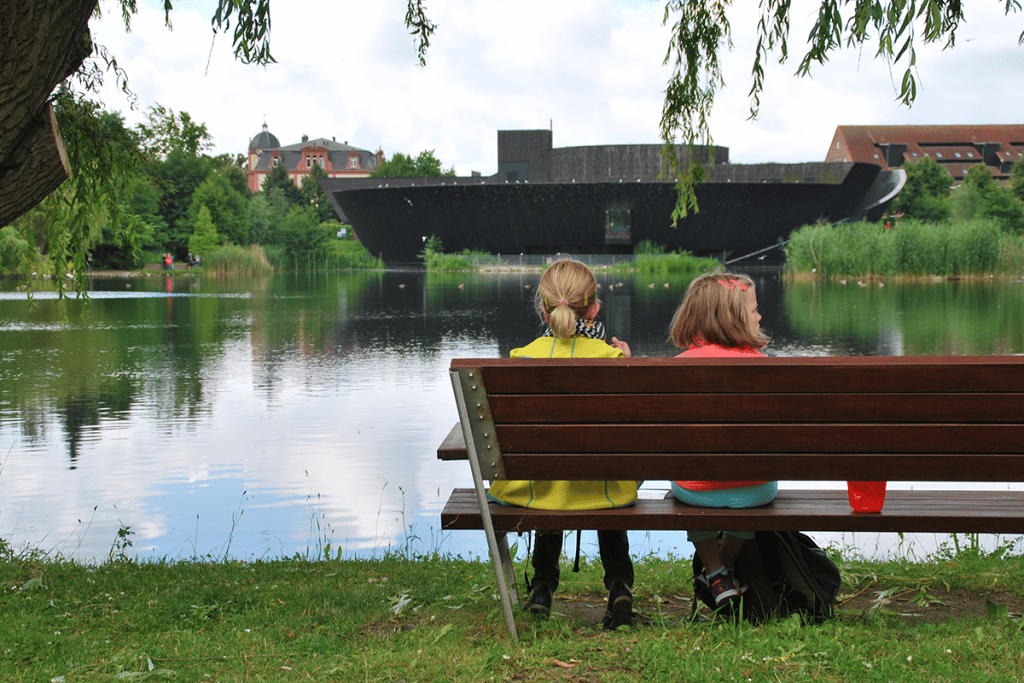 Kinder ruhen sich aus und sitzen auf einer Bank mit Blick Richtung Herrensee und das Haus der 1.000 Seen.