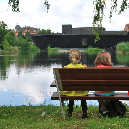 Kinder ruhen sich aus und sitzen auf einer Bank mit Blick Richtung Herrensee und das Haus der 1.000 Seen.